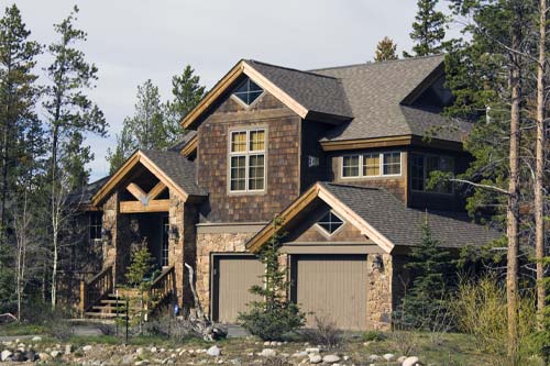Rustic two-story mountain lodge with cedar shingles and stacked stone siding, nestled in pine trees. Multi-gabled roof, front porch, and a two-car garage. Architectural photography, bright daylight.