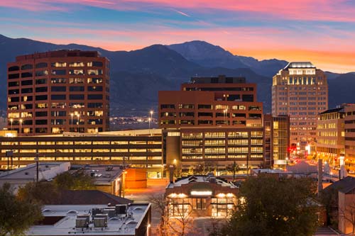 City skyline at twilight with glowing office buildings set against a dark mountain backdrop. Dramatic sunset sky with vibrant pink and orange clouds. HDR urban landscape photography.