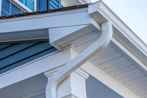 Close-up architectural shot of a clean white rain gutter and downspout on a house. Features contrasting deep navy blue vinyl siding and a white porch column. Clean lines, crisp daylight.