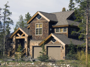 A large, rustic multi-story house featuring dark wood shingle siding, natural stone accents, a two-car garage, and a timber-framed entryway, nestled among tall pine trees.