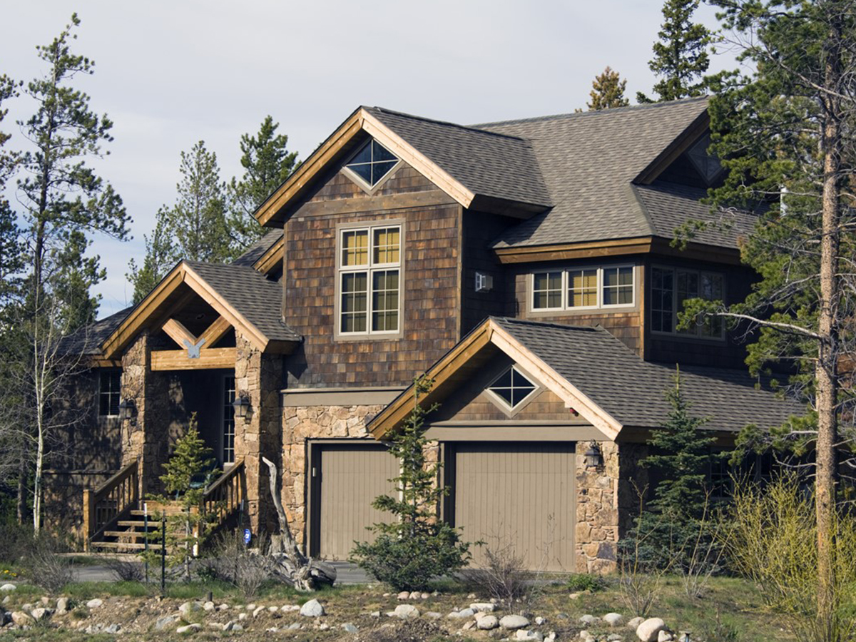 A large, rustic multi-story house featuring dark wood shingle siding, natural stone accents, a two-car garage, and a timber-framed entryway, nestled among tall pine trees.