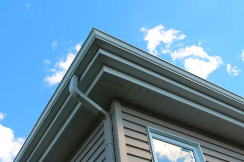 Low-angle view of a residential home's white rain gutter and downspout system against a blue sky.