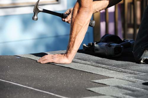 Close-up of a worker's arm using a hammer to install asphalt shingles on a roof.