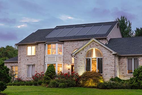 A large brick home at dusk featuring a roof-mounted solar panel array and illuminated windows.
