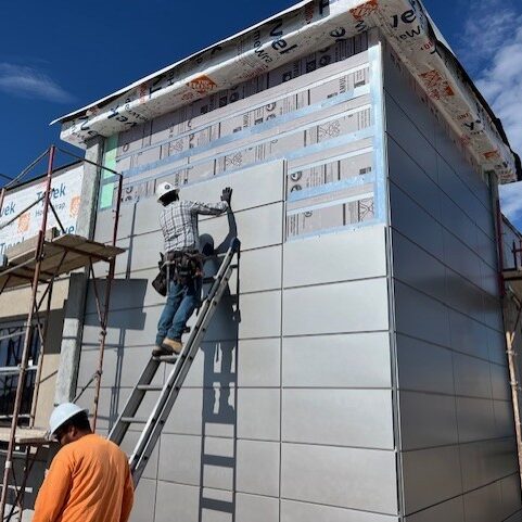 Construction worker on a ladder installing gray exterior cladding panels on a building facade.