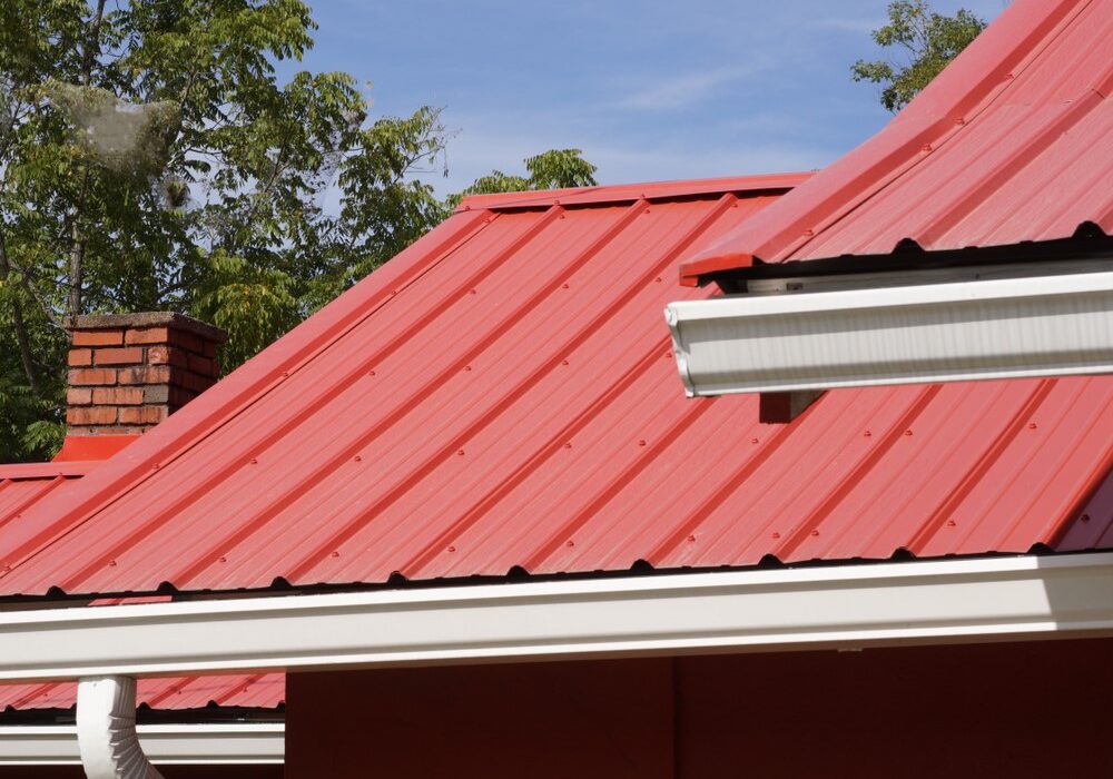 Vibrant red metal roof panels featuring white gutters, a downspout, and a brick chimney.