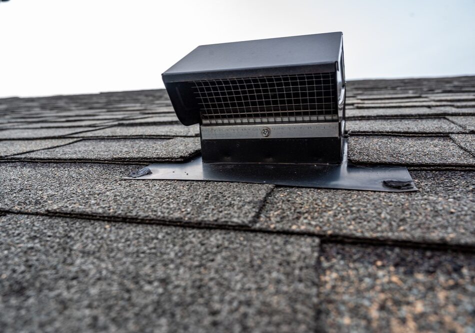 Close-up of a black metal box roof vent with a mesh screen installed on gray asphalt shingles.