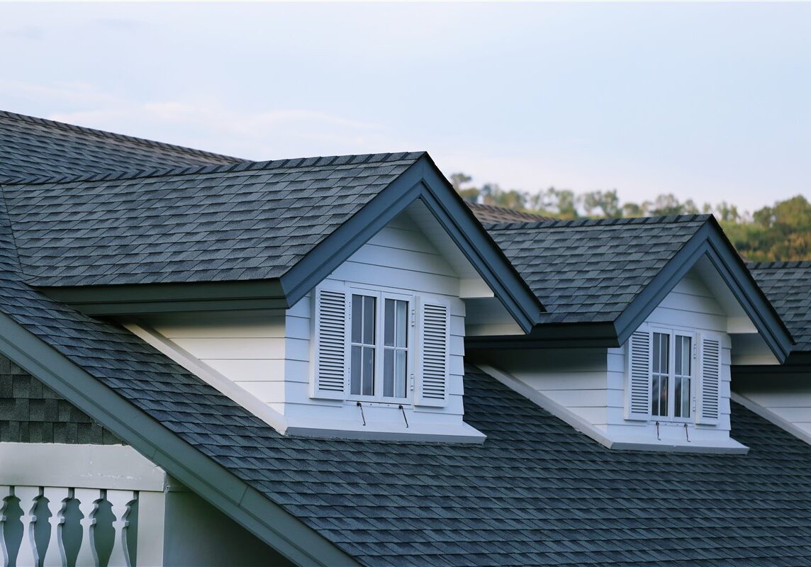 A close-up view of a residential roof featuring dark gray asphalt shingles and two white gabled dormer windows.