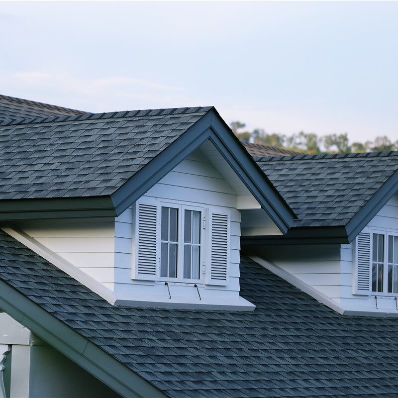 A close-up view of a residential roof featuring dark gray asphalt shingles and two white gabled dormer windows.