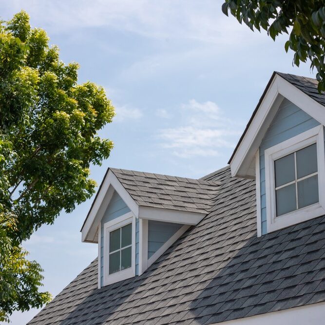 A close-up view of a residential roof with dark gray asphalt shingles and two light blue gabled dormer windows.