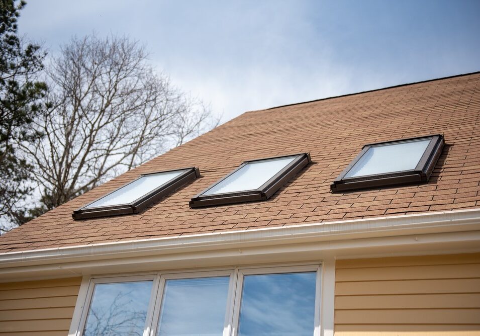 Three skylights installed in a row on a brown asphalt shingle roof above a yellow house with white gutters.