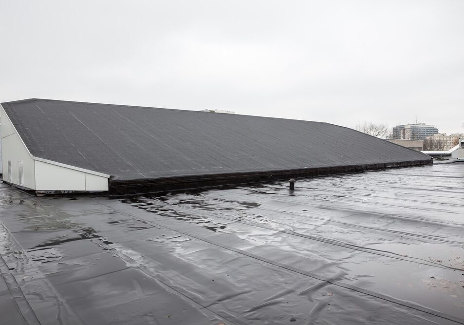 Flat commercial rooftop with a black waterproof membrane and a large sloped structure under an overcast sky.