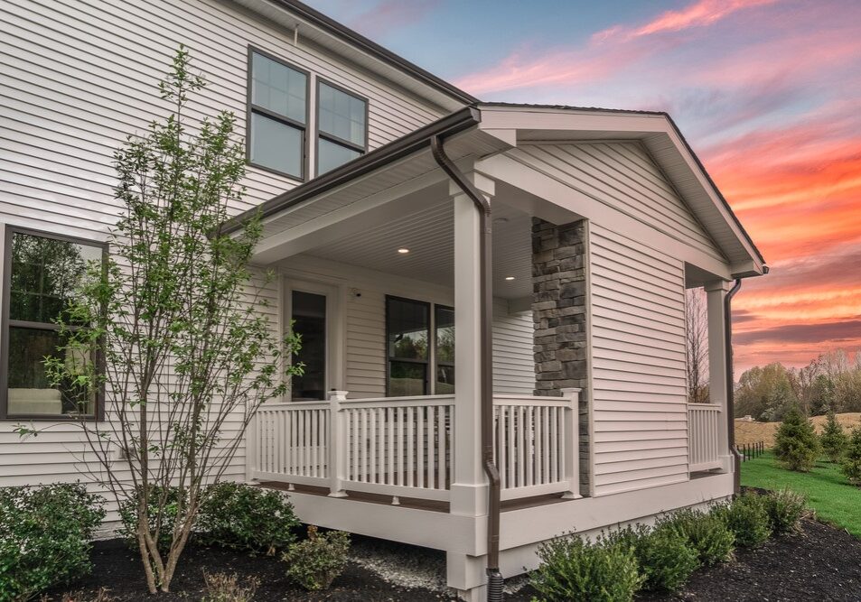 Modern residential house exterior featuring a covered porch with white railings, a stone pillar, and dark gutters against a vibrant sunset sky.