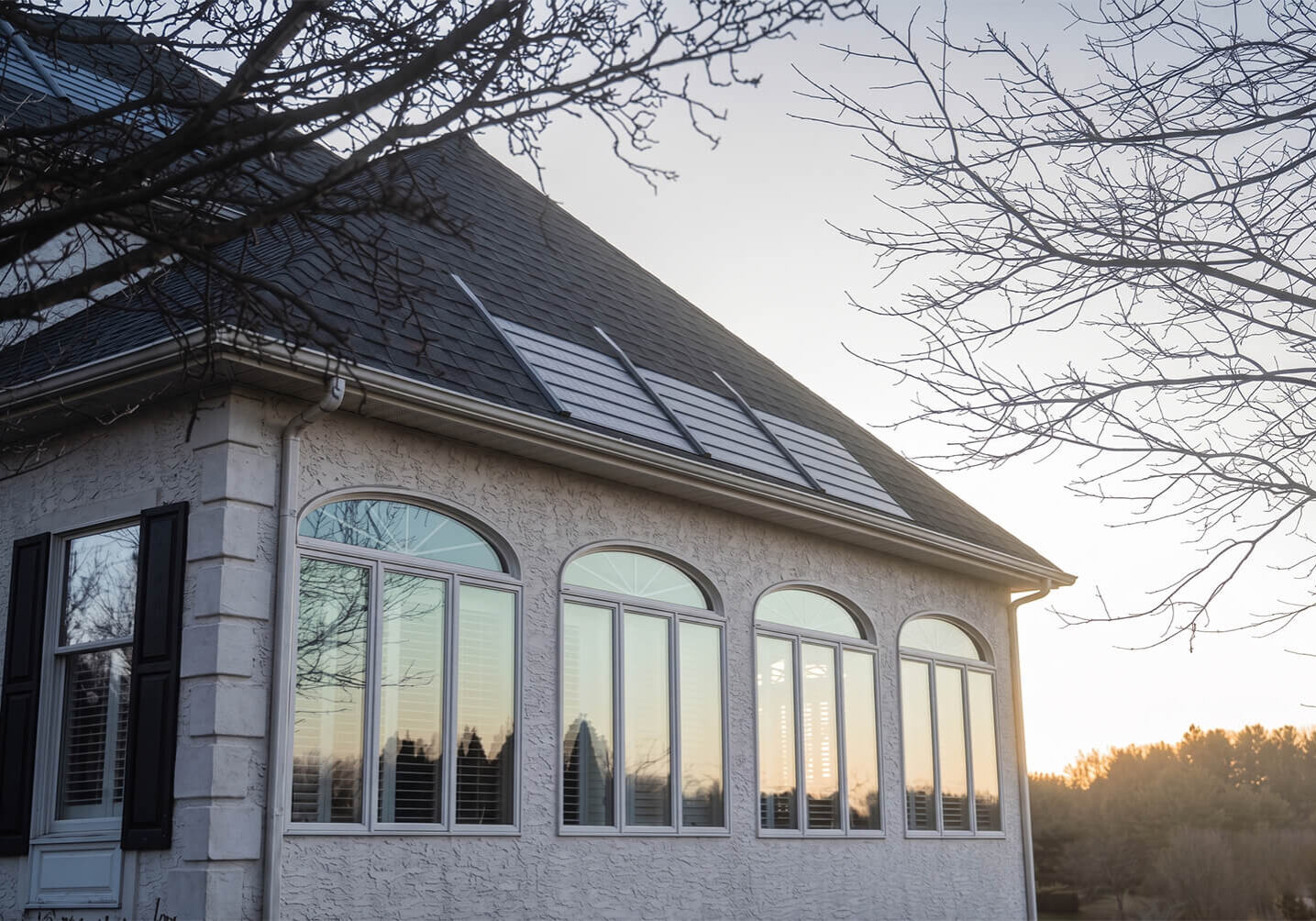 Large stucco house featuring a row of arched windows and solar panels on a dark asphalt shingle roof at sunset.