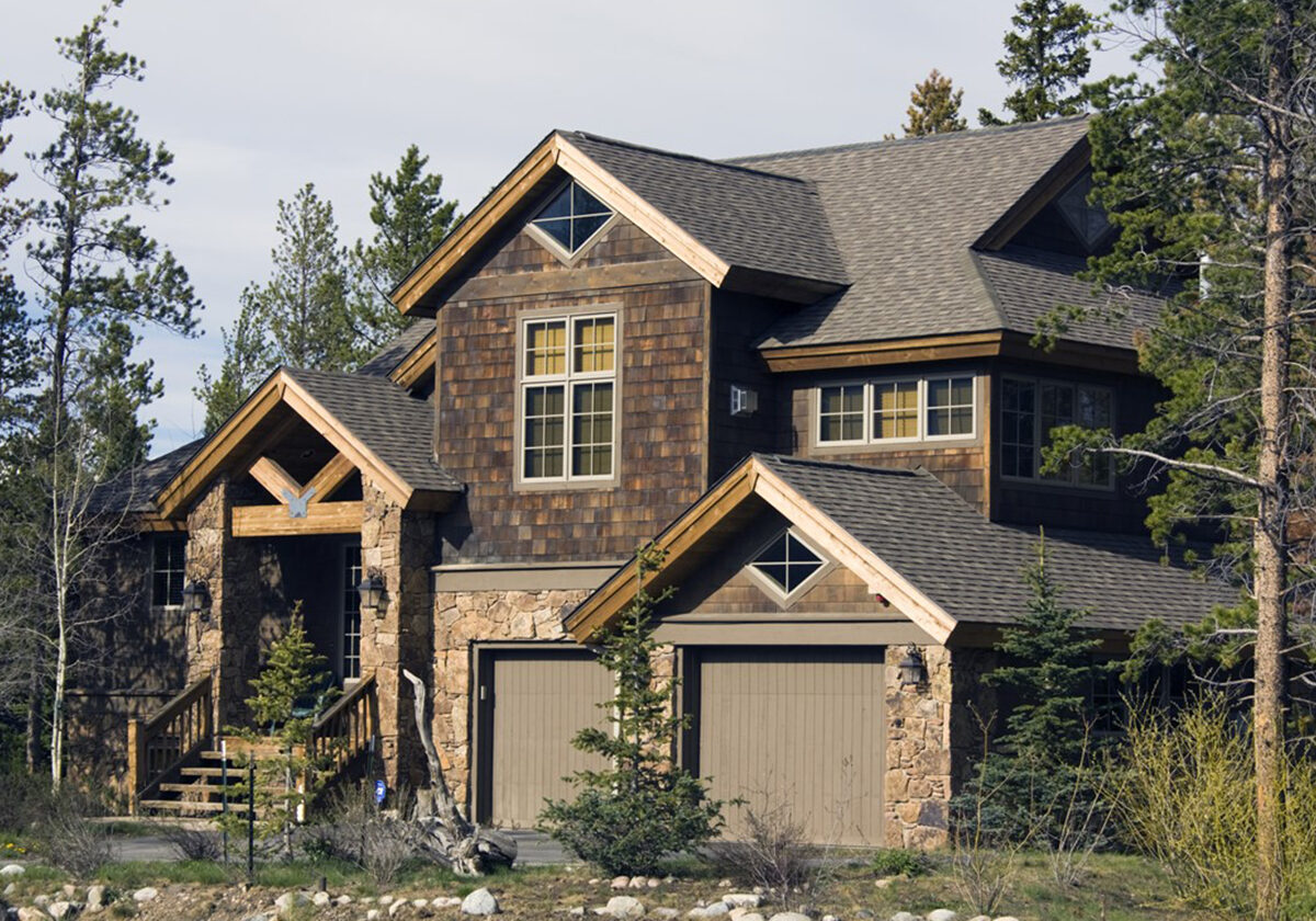A large, rustic multi-story house featuring dark wood shingle siding, natural stone accents, a two-car garage, and a timber-framed entryway, nestled among tall pine trees.