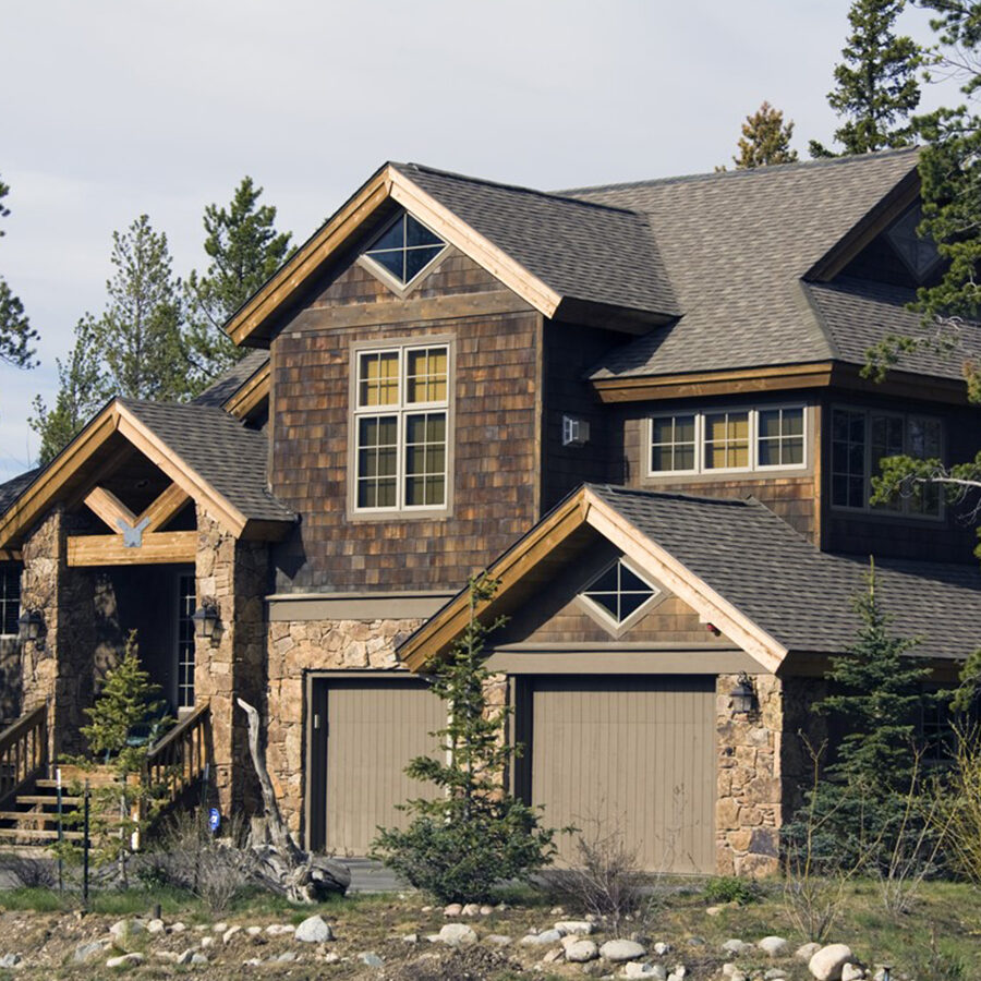 A large, rustic multi-story house featuring dark wood shingle siding, natural stone accents, a two-car garage, and a timber-framed entryway, nestled among tall pine trees.